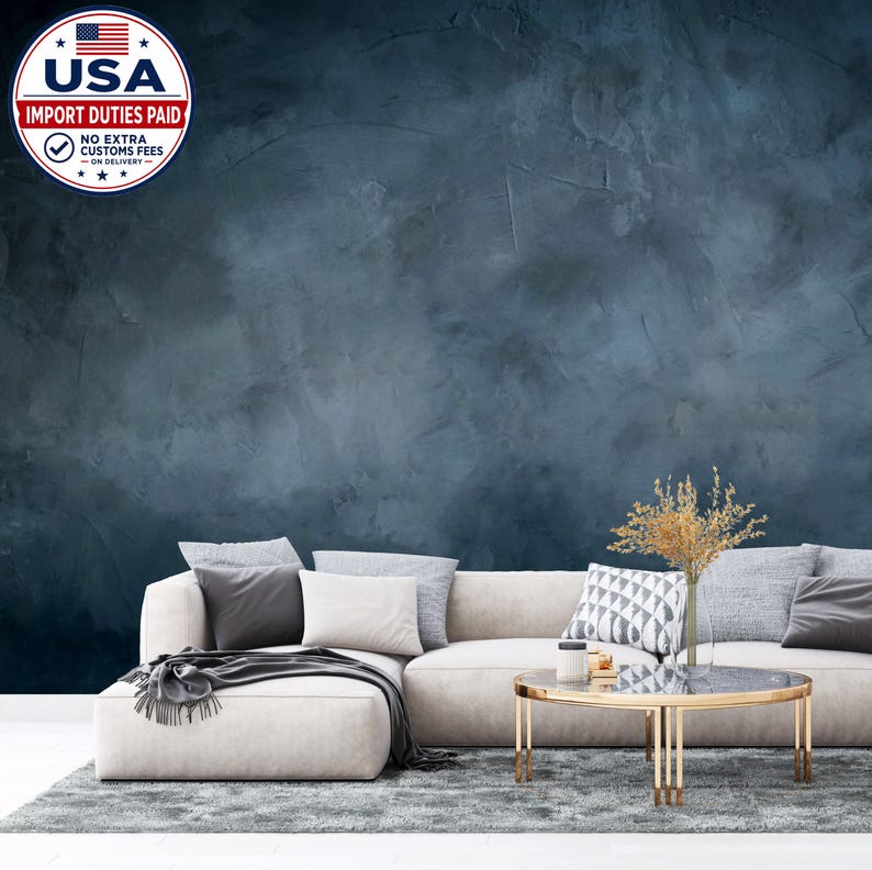May include: A dark blue wall with a textured, mottled finish behind a light beige sectional sofa. The sofa features several gray and white throw pillows. A round coffee table with a gold frame and glass top sits on a gray rug in front of the sofa.
