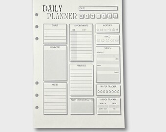 May include: A black and white daily planner with sections for goals, appointments, reminders, priorities, notes, water tracker, money tracker, and a space to write what you are grateful for.