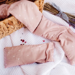 May include: Two pink fabric eye pillows with white heart patterns, lying on a white textured surface. One pillow is folded and the other is lying flat. Dried lavender flowers are in the background.