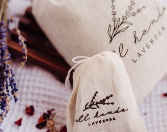 May include: Two small white fabric pouches with black drawstring closures. Both pouches have a black floral design and the text "well hands lavender" printed on them.