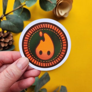 May include: Round sticker featuring a stylized orange flame design with two small black dots, set against a dark green background. The flame is encircled by a brown border with a dotted pattern and a white edge.