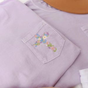 Op de afbeelding: Twee lavendelkleurige T-shirts met een geborduurd bloemenkruis op de zak. De T-shirts zijn gevouwen en liggen op een wit oppervlak.