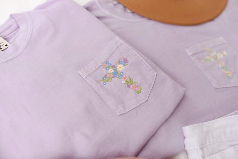 Op de afbeelding: Twee lavendelkleurige T-shirts met een geborduurd bloemenkruis op de zak. De T-shirts zijn gevouwen en liggen op een wit oppervlak.