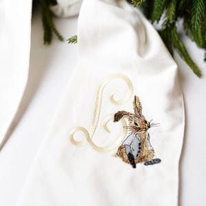 May include: White fabric with a brown rabbit and a gold embroidered letter B.