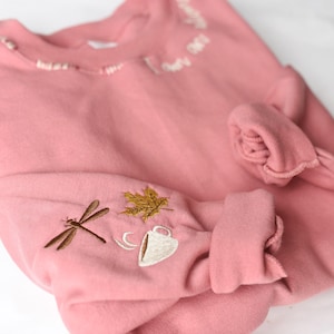 K&ouml;nnte beinhalten: Ein altrosa Sweatshirt mit gestickten Details. Am &Auml;rmel befindet sich eine Libelle, ein goldenes Blatt und eine Kaffeetasse. Der Ausschnitt hat zarte wei&szlig;e Stickereien. Die &Auml;rmel sind hochgekrempelt.