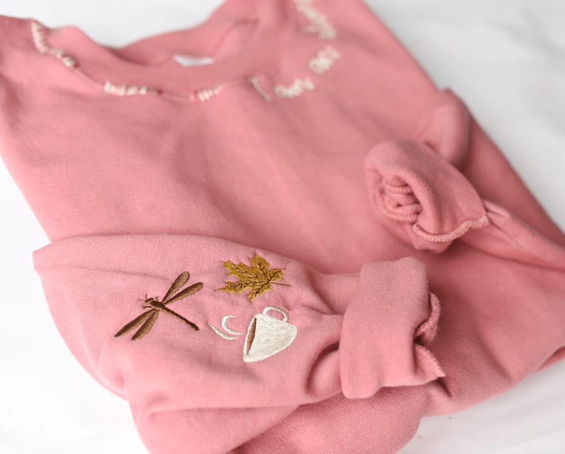K&ouml;nnte beinhalten: Ein altrosa Sweatshirt mit gestickten Details. Am &Auml;rmel befindet sich eine Libelle, ein goldenes Blatt und eine Kaffeetasse. Der Ausschnitt hat zarte wei&szlig;e Stickereien. Die &Auml;rmel sind hochgekrempelt.