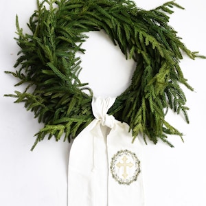 Peut inclure: Une couronne de sapin verte avec un ruban en tissu blanc noué autour. Le ruban est brodé d'une croix dorée et d'un motif de couronne.