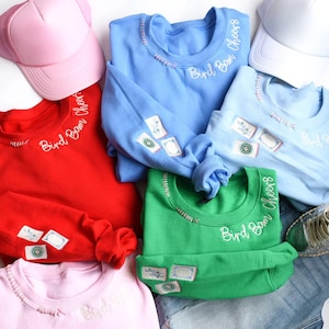 Peut inclure: Plusieurs sweatshirts colorés, rouges, bleus, verts et roses, brodés avec "Bird Bam Cheers". Les sweatshirts présentent des écussons sur les manches et sont accompagnés de casquettes de baseball et de denim.