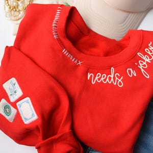 K&ouml;nnte beinhalten: Ein rotes Sweatshirt mit wei&szlig; gesticktem Text &bdquo;needs a joker&ldquo;. Das Sweatshirt hat drei gestickte Aufn&auml;her auf dem &Auml;rmel und wei&szlig;e Nahtdetails am Halsausschnitt.