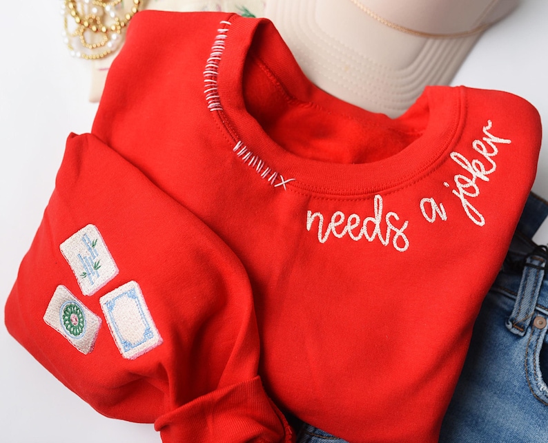 K&ouml;nnte beinhalten: Ein rotes Sweatshirt mit wei&szlig; gesticktem Text &bdquo;needs a joker&ldquo;. Das Sweatshirt hat drei gestickte Aufn&auml;her auf dem &Auml;rmel und wei&szlig;e Nahtdetails am Halsausschnitt.