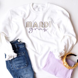 Könnte beinhalten: Weißer Pullover mit dem Text "Mardi Gras" in gelben und violetten Buchstaben. Der Pullover wird mit einer blauen Jeans und einem violetten Shirt kombiniert.
