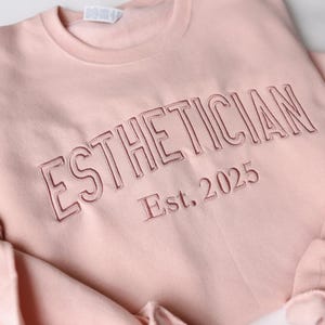 Könnte beinhalten: Hellrosa Sweatshirt mit dem Wort "ESTHETICIAN" in dunklerem Rosa auf der Brust gestickt. Darunter steht "Est. 2025". Das Sweatshirt ist aus weichem Material und hat einen Rundhalsausschnitt.