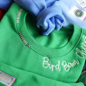 Puede incluir: Una sudadera verde con el texto bordado "Bird Bam Cheers". El cuello tiene costuras decorativas. Parches con dise&ntilde;os bordados son visibles en la sudadera. Otras sudaderas en azul y rojo est&aacute;n en el fondo.