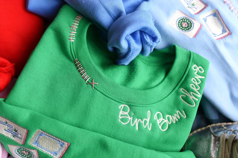 Puede incluir: Una sudadera verde con el texto bordado "Bird Bam Cheers". El cuello tiene costuras decorativas. Parches con dise&ntilde;os bordados son visibles en la sudadera. Otras sudaderas en azul y rojo est&aacute;n en el fondo.