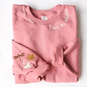 Puede incluir: Una sudadera rosa polvorienta con detalles bordados. Las palabras "I am autumn" están bordadas en blanco en el cuello. La manga izquierda presenta una libélula, una hoja de arce y una taza de té con vapor. La sudadera está doblada.