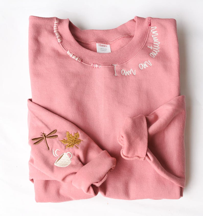 K&ouml;nnte beinhalten: Ein altrosa Sweatshirt mit gestickten Details. Die Worte "I am autumn" sind in Wei&szlig; auf den Ausschnitt gestickt. Der linke &Auml;rmel zeigt eine Libelle, ein Ahornblatt und eine Teetasse mit Dampf. Das Sweatshirt ist gefaltet.