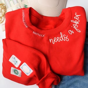 K&ouml;nnte beinhalten: Ein leuchtend rotes Sweatshirt mit dem gestickten Schriftzug "needs a joker" in wei&szlig;er Schrift. Das Sweatshirt hat dekorative N&auml;hte am Halsausschnitt und Aufn&auml;her am &Auml;rmel. Das Kleidungsst&uuml;ck wird mit einer blauen Jeans kombiniert.