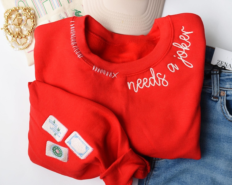 K&ouml;nnte beinhalten: Ein leuchtend rotes Sweatshirt mit dem gestickten Schriftzug "needs a joker" in wei&szlig;er Schrift. Das Sweatshirt hat dekorative N&auml;hte am Halsausschnitt und Aufn&auml;her am &Auml;rmel. Das Kleidungsst&uuml;ck wird mit einer blauen Jeans kombiniert.