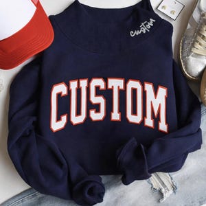 Puede incluir: Sudadera azul marino con la palabra "CUSTOM" en letras blancas grandes con ribete naranja en la parte delantera. La palabra "custom" también está bordada en cursiva en el cuello. Una gorra de camionero roja y blanca es visible en la esquina superior izquierda.