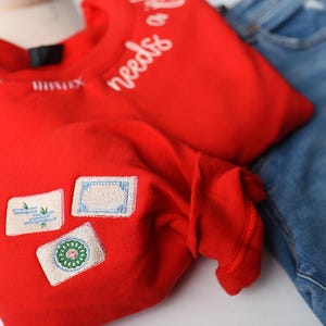 K&ouml;nnte beinhalten: Ein leuchtend rotes Langarmshirt mit dem wei&szlig;en Schriftzug "needs of" auf der Schulter. Das Shirt hat drei gestickte Aufn&auml;her auf der Vorderseite. Eine blaue Jeans ist im Hintergrund zu sehen.