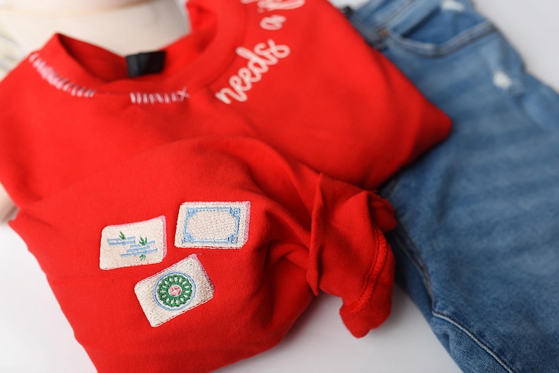 K&ouml;nnte beinhalten: Ein leuchtend rotes Langarmshirt mit dem wei&szlig;en Schriftzug "needs of" auf der Schulter. Das Shirt hat drei gestickte Aufn&auml;her auf der Vorderseite. Eine blaue Jeans ist im Hintergrund zu sehen.