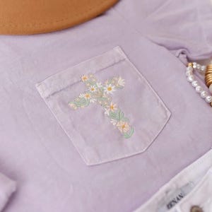 Op de afbeelding: Een lavendelkleurig T-shirt met een geborduurd bloemenkruis op de zak. Het shirt wordt gecombineerd met een witte spijkerbroek en een gouden kralenarmband.