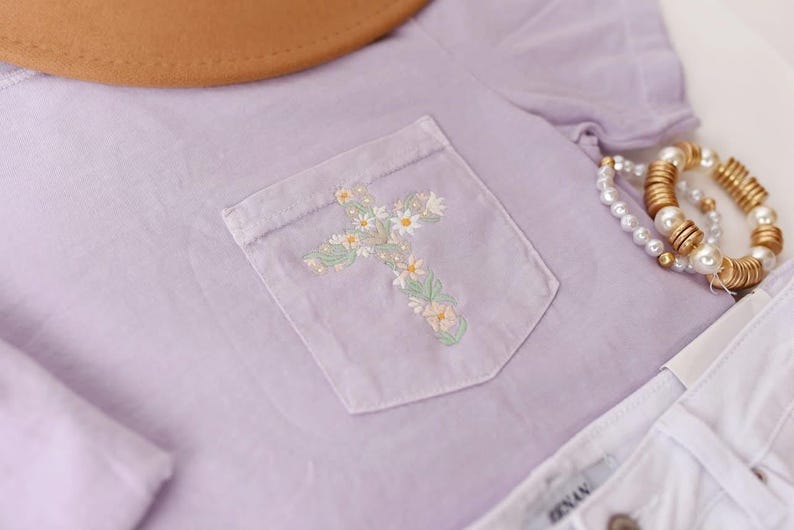 Op de afbeelding: Een lavendelkleurig T-shirt met een geborduurd bloemenkruis op de zak. Het shirt wordt gecombineerd met een witte spijkerbroek en een gouden kralenarmband.
