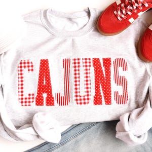 May include: A light grey crewneck sweatshirt featuring the word CAJUNS in large, block-style letters. Each letter is filled with alternating red and white patterns, including gingham, polka dots, and vertical stripes. A white cap and red trainers.