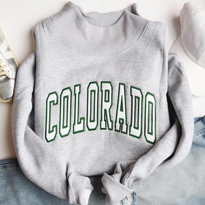 Könnte beinhalten: Ein hellgraues Langarm-Sweatshirt mit Stehkragen. Das Wort COLORADO ist in einer fetten, geschwungenen Schriftart mit weißen Buchstaben und einer dunkelgrünen Umrandung auf die Brust gestickt, was ein Design im College-Stil erzeugt.