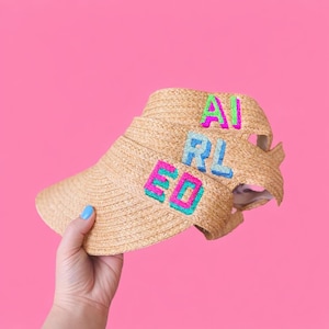 Può includere: Visiera di paglia con la scritta "AIRED" ricamata in lettere colorate. Le lettere sono rosa, blu e verdi. Lo sfondo è rosa acceso.