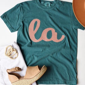 May include: A teal blue t-shirt with a pink "la" graphic on the front. The shirt is made of a soft, comfortable material and has a relaxed fit.