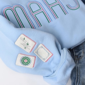 Pode incluir: Camisola azul clara com a palavra "MAHS" bordada em verde, rosa e azul. A manga apresenta tr&ecirc;s patches bordados com v&aacute;rios desenhos. A camisola &eacute; combinada com cal&ccedil;as de ganga azuis.
