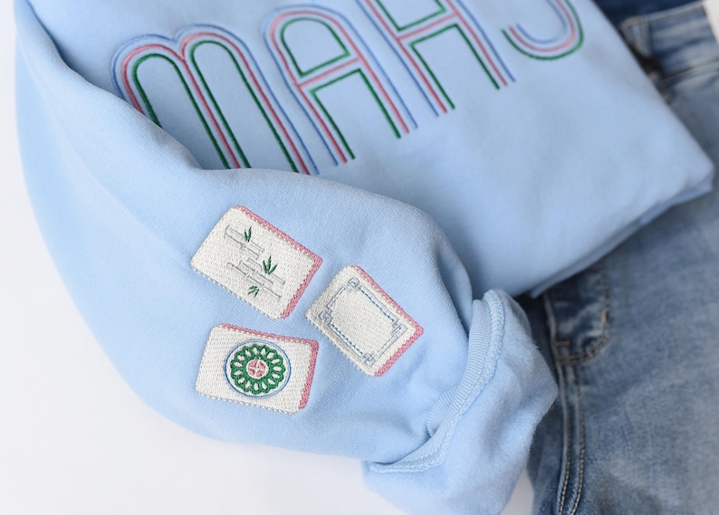 Pode incluir: Camisola azul clara com a palavra "MAHS" bordada em verde, rosa e azul. A manga apresenta tr&ecirc;s patches bordados com v&aacute;rios desenhos. A camisola &eacute; combinada com cal&ccedil;as de ganga azuis.