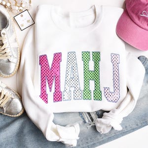 May include: White sweatshirt with the letters "MAHJ" in pink, green, and purple sequined polka dots. The sweatshirt is folded on top of a pair of blue jeans. A pink baseball cap and silver sneakers are also in the image.