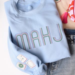 Op de afbeelding: Een lichtblauw sweatshirt met het woord "MAHJ" geborduurd in groen, rood en blauw. De mouw heeft geborduurde patches. Het sweatshirt wordt gecombineerd met een spijkerbroek en een kleine roze en witte tas.