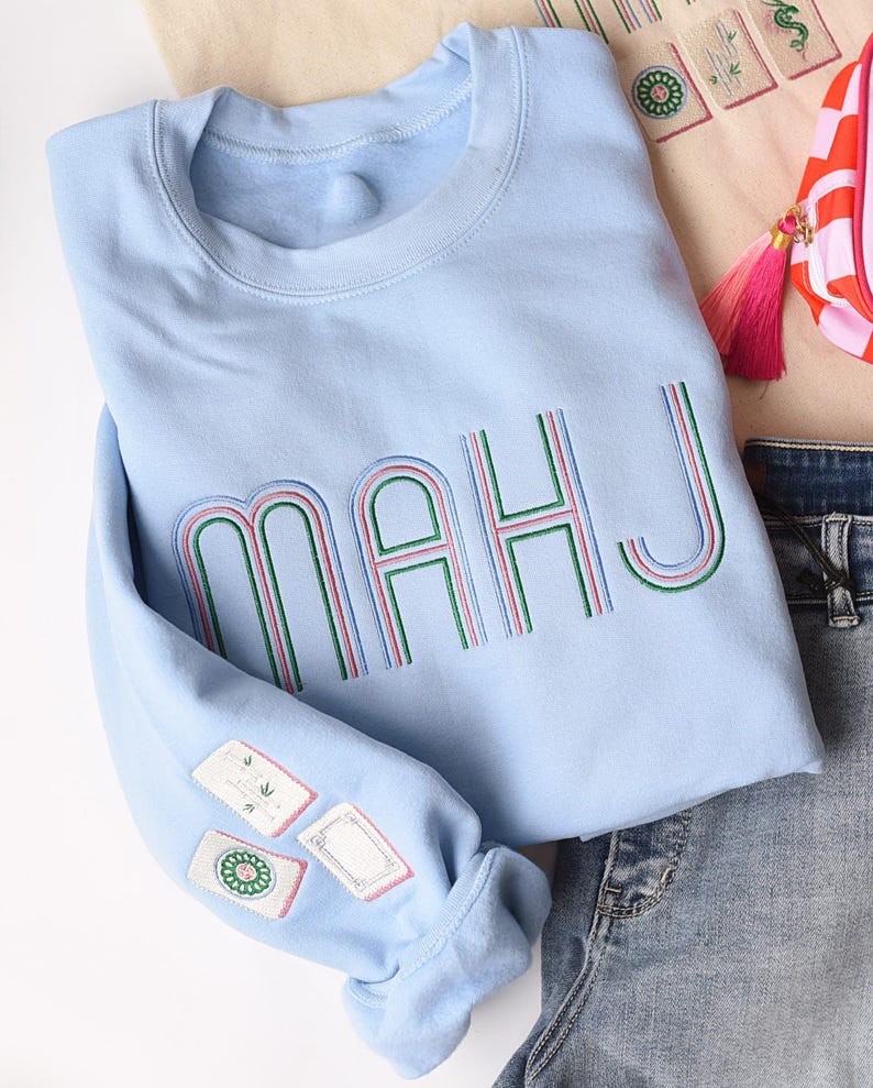 Pode incluir: Camisola azul clara com a palavra "MAHJ" bordada em verde, vermelho e azul. A manga tem patches bordados. A camisola &eacute; combinada com jeans e uma pequena bolsa rosa e branca.