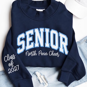 Könnte beinhalten: Ein marineblaues Sweatshirt mit Rundhalsausschnitt und dem Wort SENIOR in großen weißen und hellblauen Blockbuchstaben auf der Brust. Darunter ist der Text North Penn Cheer in Weiß gestickt. Der Ärmel zeigt Class of 2027 in Weiß.
