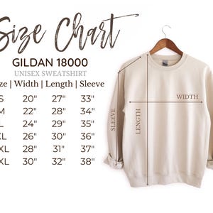 Puede incluir: Una sudadera unisex Gildan 18000 de color crema cuelga de una percha de madera. La imagen incluye una tabla de tallas con medidas en cm para el ancho, el largo y el largo de la manga, desde la talla S hasta la 3XL.