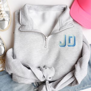 Könnte beinhalten: Hellgraues Sweatshirt mit Viertelreißverschluss und den gestickten Buchstaben "JD" in Hellblau und Blau. Das Sweatshirt ist gefaltet, mit einem silbernen Reißverschluss und einem weißen Innenfutter. Eine rosa Baseballkappe, silberne Turnschuhe und Goldschmuck sind ebenfalls zu sehen.