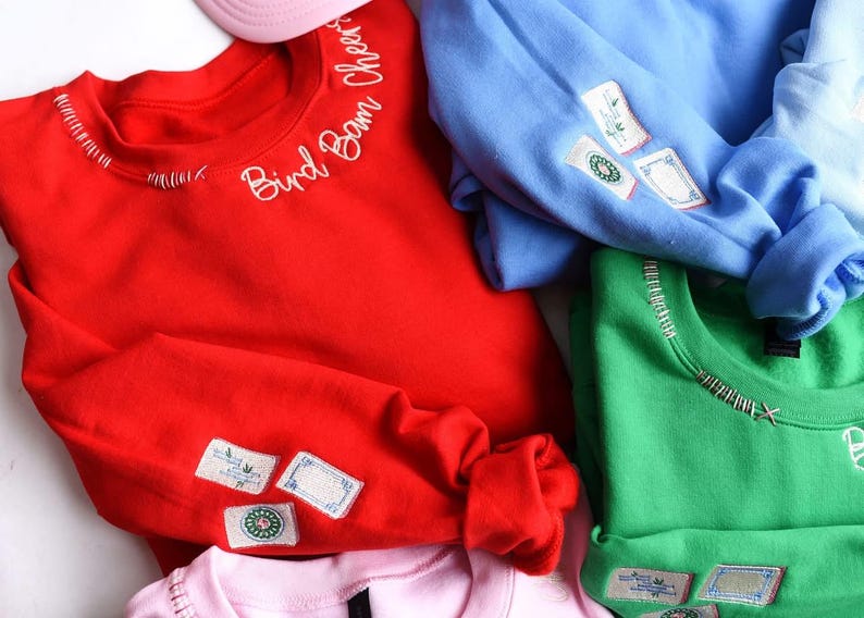 Puede incluir: Una colecci&oacute;n de sudaderas coloridas en rojo, azul, verde y rosa. Cada sudadera tiene texto bordado y parches en las mangas. La sudadera roja tiene bordado "Bird Bam Cheers".