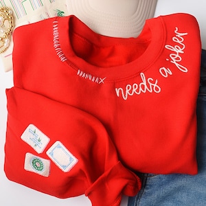 K&ouml;nnte beinhalten: Ein leuchtend rotes Sweatshirt mit dem gestickten Schriftzug "needs a joker" in wei&szlig;er Schrift. Der Halsausschnitt hat dekorative N&auml;hte, und der &Auml;rmel hat drei gestickte Aufn&auml;her mit wei&szlig;en Hintergr&uuml;nden und gr&uuml;nen und blauen Designs.