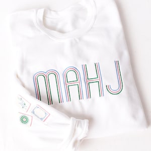 Pode incluir: Camisola branca com as letras "MAHJ" bordadas em verde, rosa e azul. A manga apresenta desenhos bordados. A camisola est&aacute; dobrada, com a manga parcialmente vis&iacute;vel.