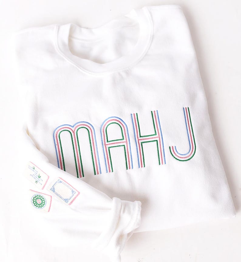 Pode incluir: Camisola branca com as letras "MAHJ" bordadas em verde, rosa e azul. A manga apresenta desenhos bordados. A camisola est&aacute; dobrada, com a manga parcialmente vis&iacute;vel.