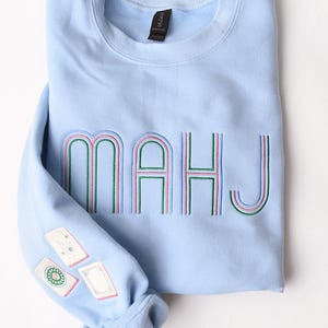 Pode incluir: Camisola azul clara com a palavra "MAHJ" bordada em verde, rosa e azul. A manga apresenta patches bordados. A camisola est&aacute; dobrada cuidadosamente sobre uma superf&iacute;cie branca.
