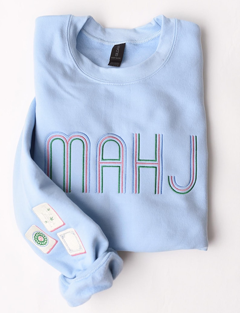 Pode incluir: Camisola azul clara com a palavra "MAHJ" bordada em verde, rosa e azul. A manga apresenta patches bordados. A camisola est&aacute; dobrada cuidadosamente sobre uma superf&iacute;cie branca.