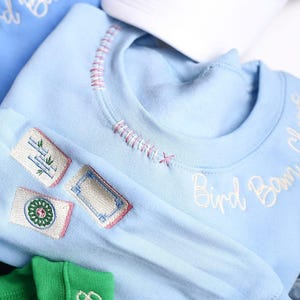 Puede incluir: Sudadera azul claro con detalles bordados. Presenta tres parches con dise&ntilde;os bordados y las palabras "Bird Boom Cheers" en escritura blanca. El cuello tiene costuras decorativas.