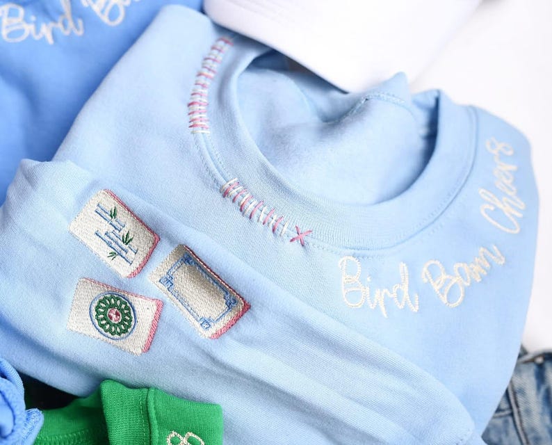 Puede incluir: Sudadera azul claro con detalles bordados. Presenta tres parches con dise&ntilde;os bordados y las palabras "Bird Boom Cheers" en escritura blanca. El cuello tiene costuras decorativas.