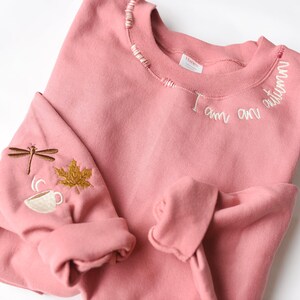 K&ouml;nnte beinhalten: Ein altrosa Sweatshirt mit gestickten Details. Der Text "I am an autumn" ist um den Halsausschnitt gestickt. Der linke &Auml;rmel zeigt eine Libelle, ein Ahornblatt und eine Kaffeetasse, alle in komplement&auml;ren Farben gestickt.