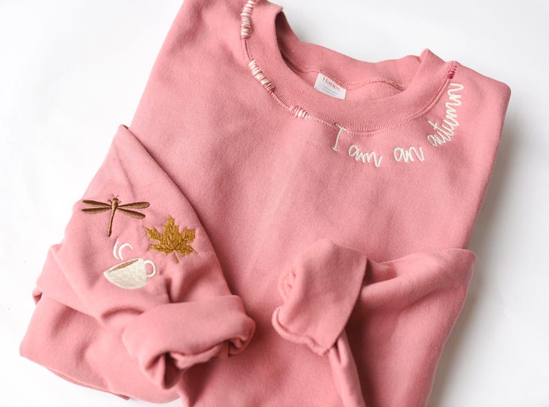 K&ouml;nnte beinhalten: Ein altrosa Sweatshirt mit gestickten Details. Der Text "I am an autumn" ist um den Halsausschnitt gestickt. Der linke &Auml;rmel zeigt eine Libelle, ein Ahornblatt und eine Kaffeetasse, alle in komplement&auml;ren Farben gestickt.