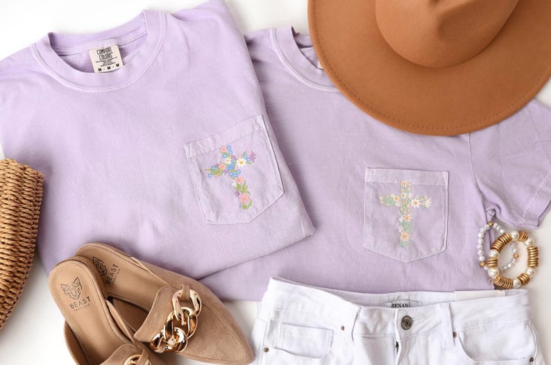 Op de afbeelding: Twee lavendelkleurige T-shirts met een geborduurd bloemenkruis op de zak. De T-shirts liggen op een wit oppervlak met een bruine hoed, een strooien tas en een paar bruine schoenen.